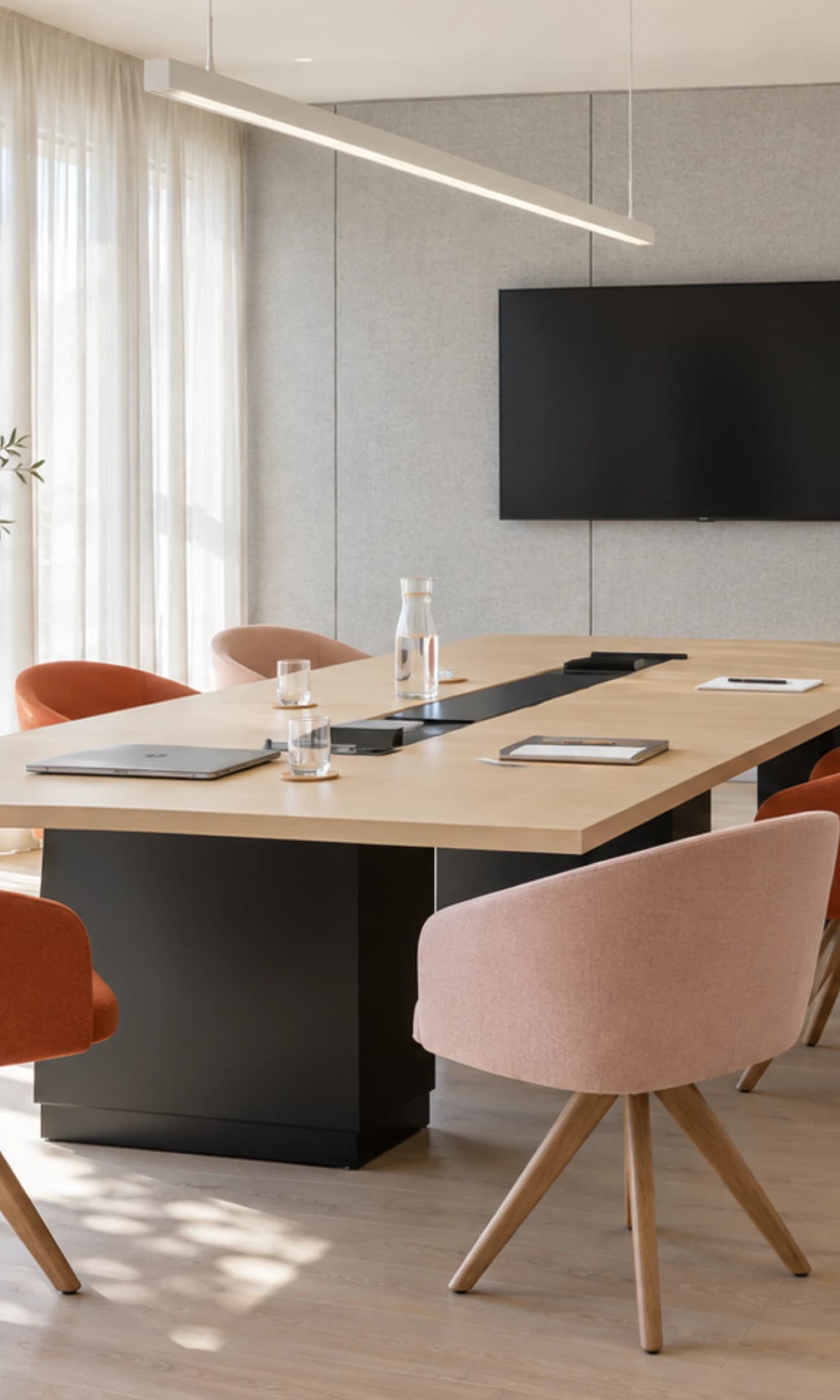 Sala de juntas con mesa de madera clara, sillas en terracota y rosa, y muro de paneles grises